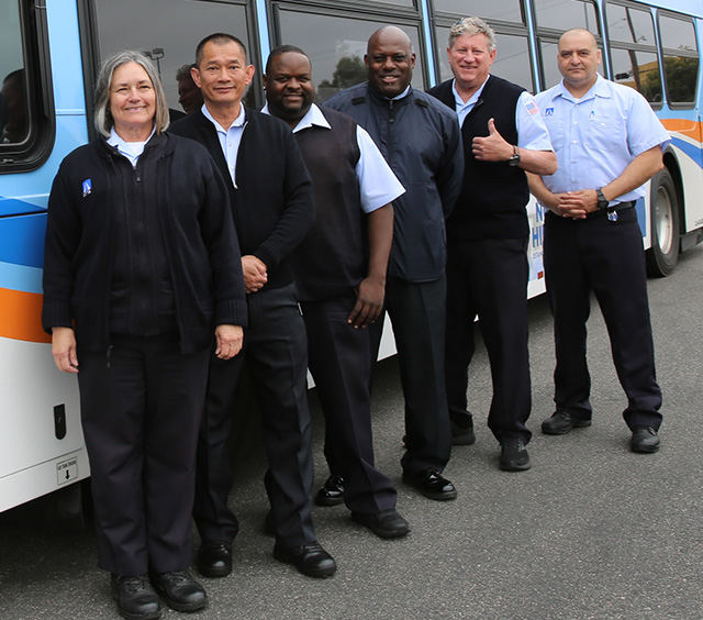 A group of smiling OC Bus drivers stand in a row in front of an OC Bus