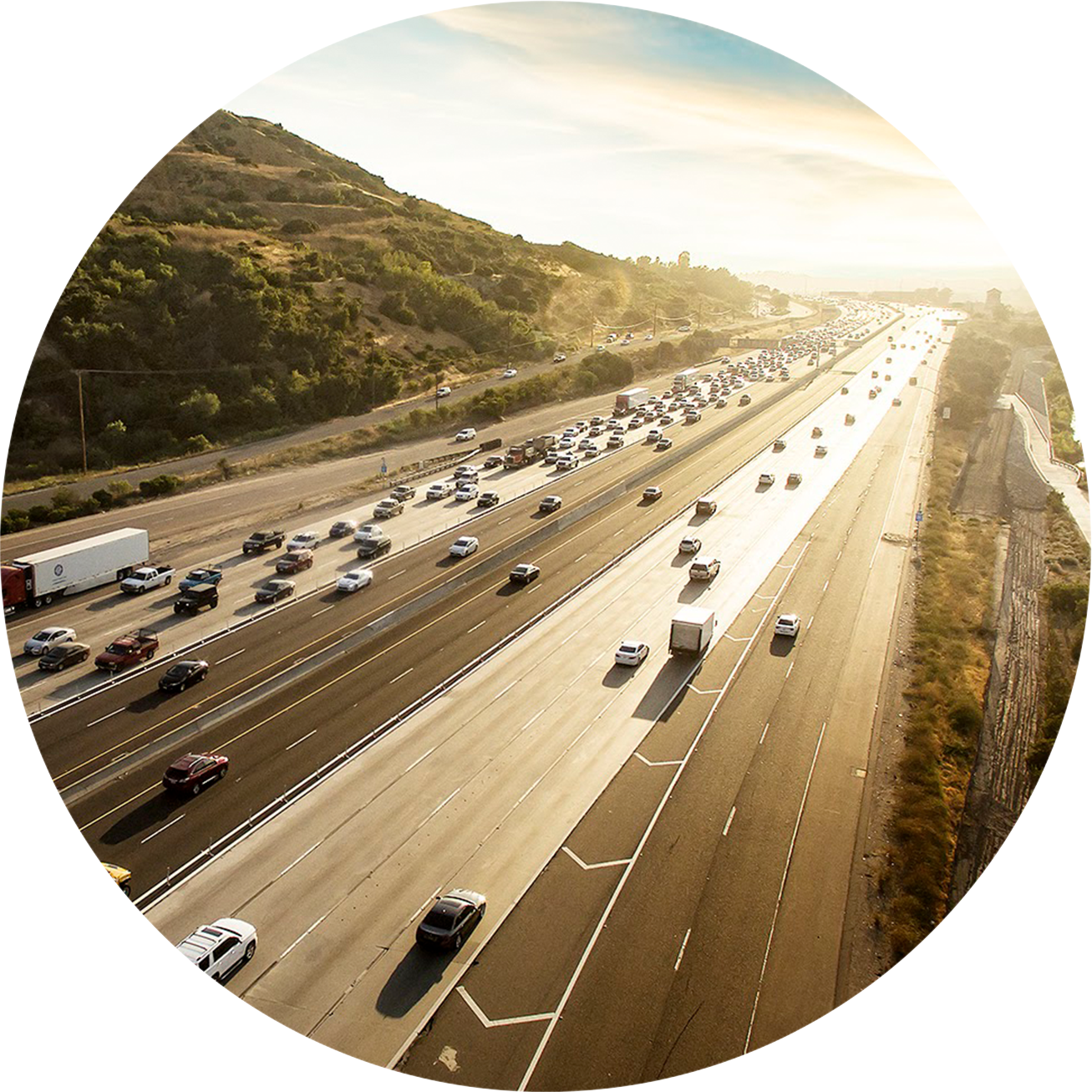 a stretch of freeway with traffic during sunrise with a shrubby hill to the left and flat shrub land to the right